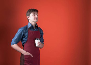 kırmızı önlük koyu saçlı yakışıklı barista yan görünümü kahve ile kağıt fincan tutan ve mercan rengi arka plan üzerinde uzağa bakıyor
