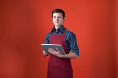 bordo önlük yakışıklı barista kamera bakarak ve kopya alanı ile turuncu arka plan üzerinde dijital tablet kullanarak