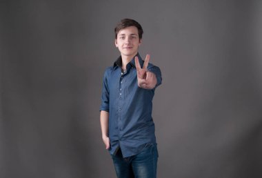 mavi yakışıklı genç adam kameraya bakıyor ve kopya alanı ile gri arka plan üzerinde el ile zafer sembolü gösteren