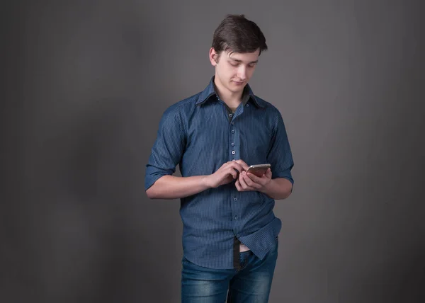 青いシャツを着た自信のあるハンサムな若者が コピースペースを持つ灰色の背景にスマートフォンを保持し 使用しています — ストック写真