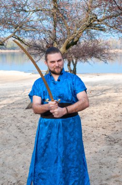 mavi kimono içinde kendine güvenen yakışıklı adam başında topuz kılıç tutan ve ağaçlar ve su yakınındaki plajda kamera bakıyor