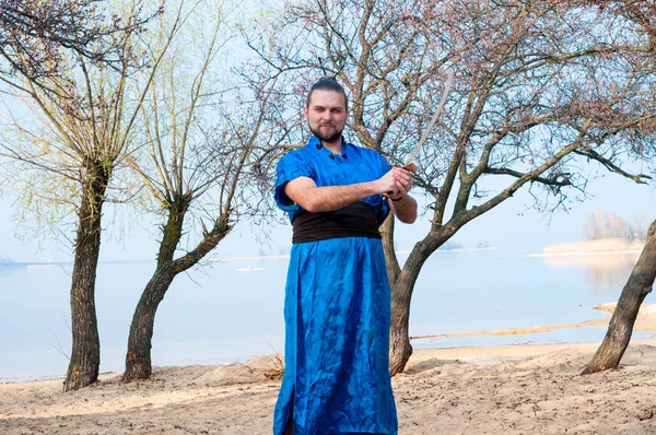 handsome man in blue kimono with belt, bun and sticks on head holding sword and looking at camera on sandy beach near trees and river