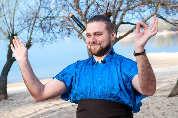 stock image overweight handsome smiling samurai in blue kimono, standing, laughing and showing meditation on beach near trees and water