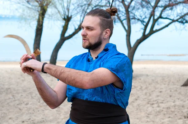 enthusiastic man in blue kimono, bun and sticks on head training with sword and looking away on sandy river beach in front of trees and water