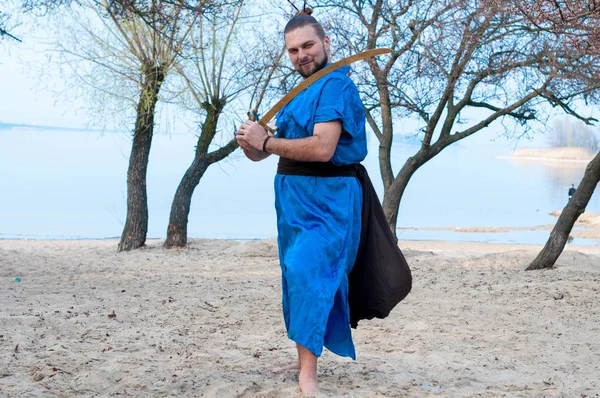 man in blue kimono, bun and sticks on head training with sword and looking away on sandy river beach in front of trees and water