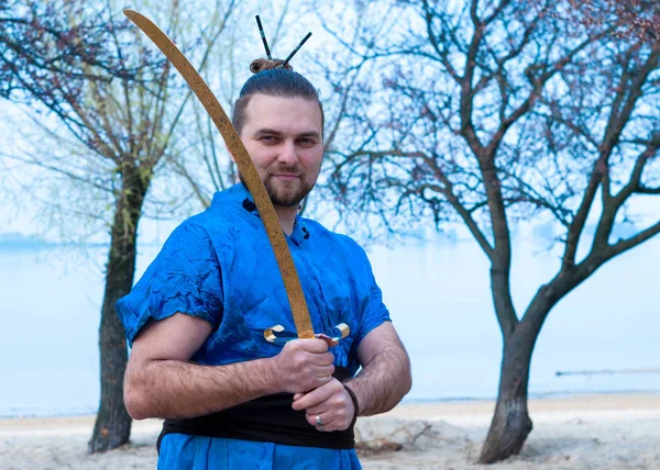 handsome man in blue kimono, bun and sticks on head holding sword and looking at camera in front of trees and river