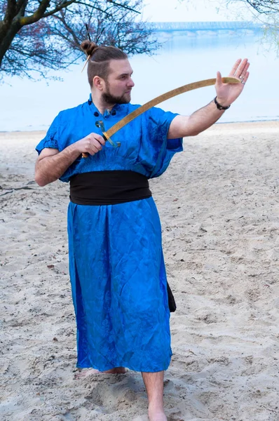 handsome man in blue kimono with belt, bun and sticks on head holding sword and looking away on sandy beach near trees and river