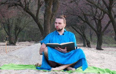 makyaj ile yakışıklı sakallı adam, mavi kimono oturan baş topuz, büyük kitap tutan ve ağaçların yakınındaki kumlu plajda uzağa bakıyor