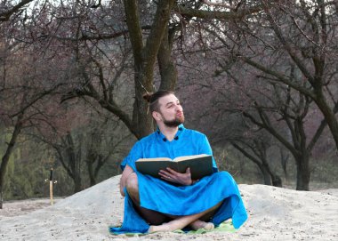 ağaçların yakınındaki kumlu plajda büyük bir kitap ile oturan mavi kimono zor sakallı adam