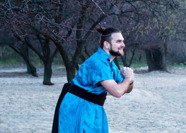 mavi kimono ile duran gülen yakışıklı sakallı adam yan görünümü, ağaçların önünde kumlu plajda uzağa bakıyor