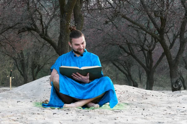 surprised bearded man in blue kimono sitting with large book on sandy beach near trees