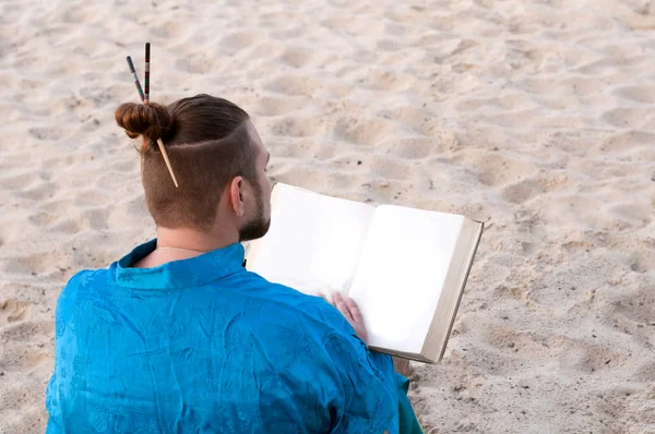 back view of bearded man with bun on head in blue kimono sitting, holding large book on sandy beach