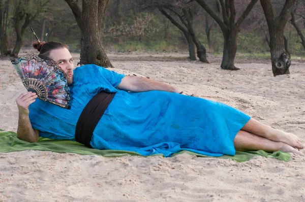 embarrassed transsexual handsome man with make up, hair bun lying on sand in blue kimono, holding hand fan and looking at camera on rug
