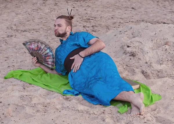 transsexual handsome man with make up, hair bun lying on sand in blue kimono, giving kiss, holding hand fan and looking away on rug