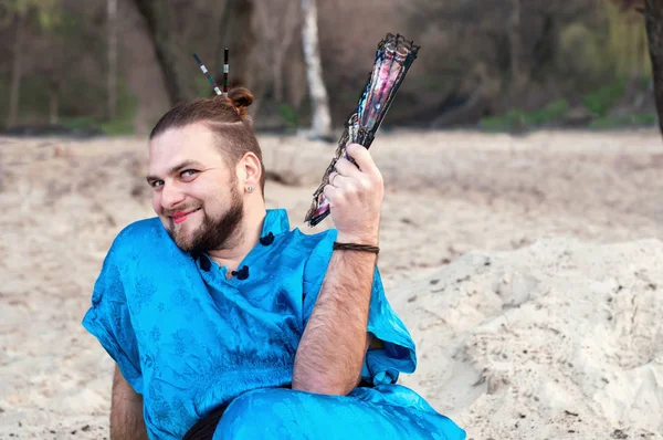 transsexual handsome man with make up, hair bun sitting on sand in blue kimono, smiling and holding hand fan in front of trees