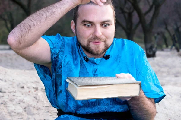 青い着物を着た集中したハンサムな男が 木々の前の浜辺の本に砂を注ぐ — ストック写真