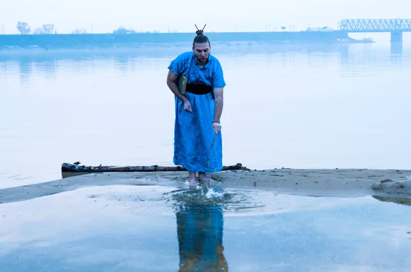 fooling around bearded man in blue kimono holding book, standing on river bank, splashing water with wooden stick in front of bridge and looking on reflection