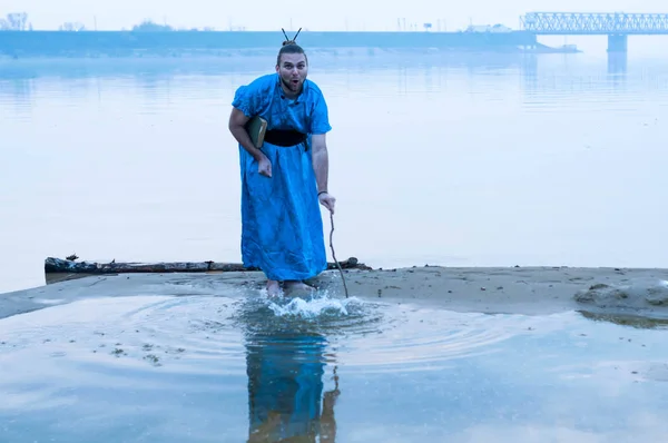 tricky man in blue kimono with hair bun splashing water with wooden stick on river bank