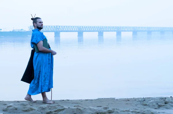 overweight bearded man in blue kimono with book on river bank in fog looking away in front of bridge