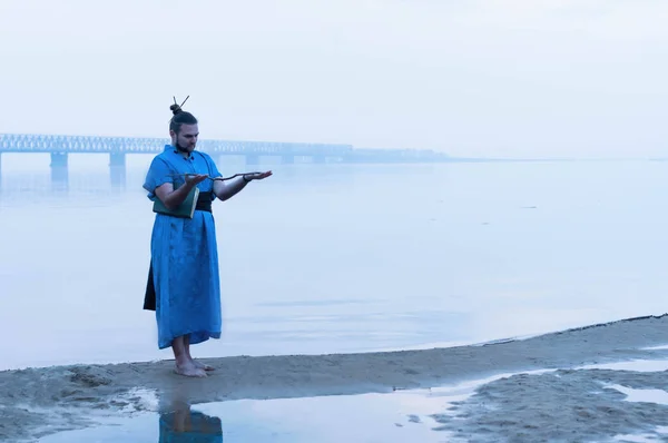 overweight bearded man in blue kimono standing on river bank in fog looking at wooden stick in front of bridge