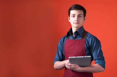 mavi gömlek ve bordo önlük dijital tablet tutan koyu saçlı genç barista ve mercan arka plan önünde kamera bakıyor