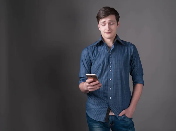 confused and scared young man with dark hair in blue shirt, holding hand in pocket and looking at smartphone in front of gray background