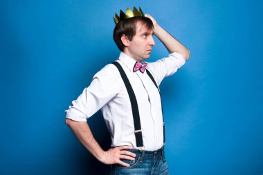 pembe gömlek koyu saçlı yakışıklı adam yan görünümü, papyon ve siyah askı kalça el tutan, altın parlak taç düzeltme ve kopya alanı ile mavi arka plan üzerinde uzağa bakıyor