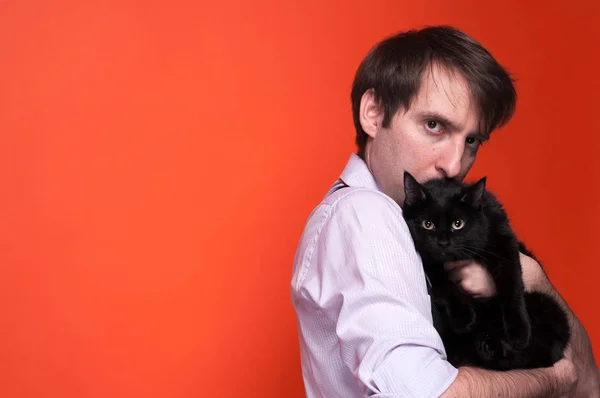 ピンクのシャツを着た心配した男の側面図は かわいい黒猫を抱きしめ コピースペースを持つサンゴの背景にカメラでくっついて 巻いた袖を持って抱きしめる — ストック写真