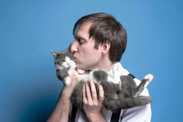 handsome man with closed eyes in shirt and black suspender holding and kissing cute gray and white cat muzzle on blue background