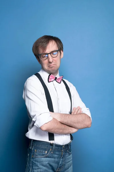 tricky man with crossed arms in pink shirt with rolled up sleeves, bow tie, black suspender and glasses grimacing and looking down on blue background