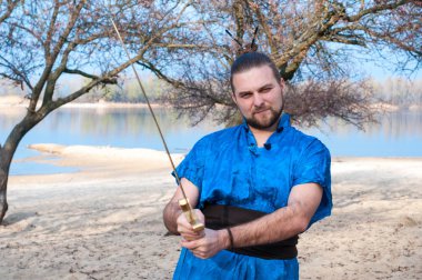 altın kılıç tutan mavi kimono başında topuz ile yakışıklı sakallı adam ve nehir yakınındaki ağaçların önünde kamera bakıyor