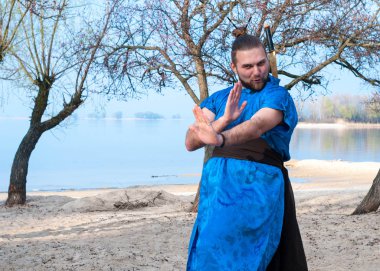 kung fu teknikleri gösteren ve ağaçlar ve nehir yakınındaki kumlu plajda kameraya bakarak çapraz elleri ile mavi kimono baş topuz ile gülümseyen ve komik yakışıklı sakallı adam 
