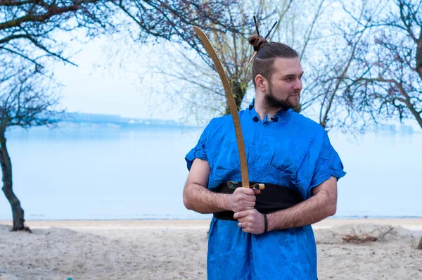 handsome bearded man with bun on head in blue kimono holding golden sword and looking away on beach in front of trees near river