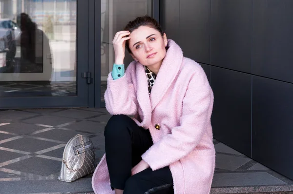 dreaming woman with bundled hair in pink coat and black pants sitting on stairs, straightens hair with hand and looking away near building