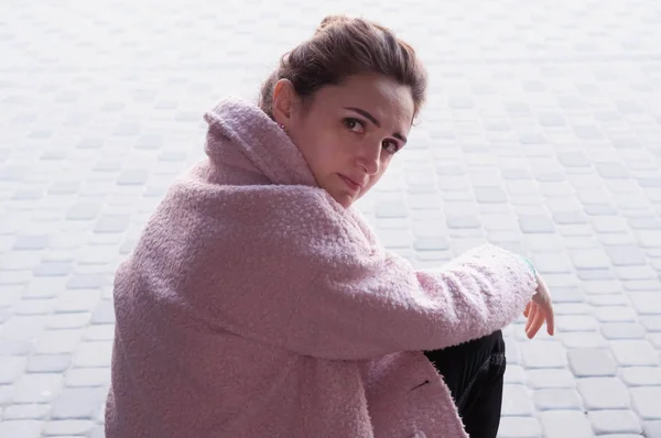 side view of woman with bundled hair in pink coat and black pants sitting on stairs and crying near building and brick sidewalk