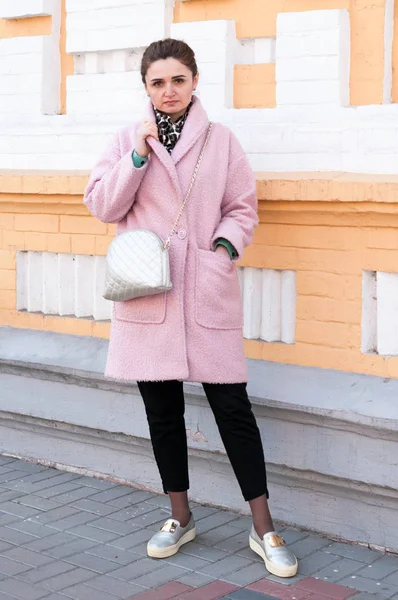 ピンクのコートと黒いズボンの美しい自信に満ちた女性が立って 建物のレンガのファサードの近くにカメラを見て — ストック写真