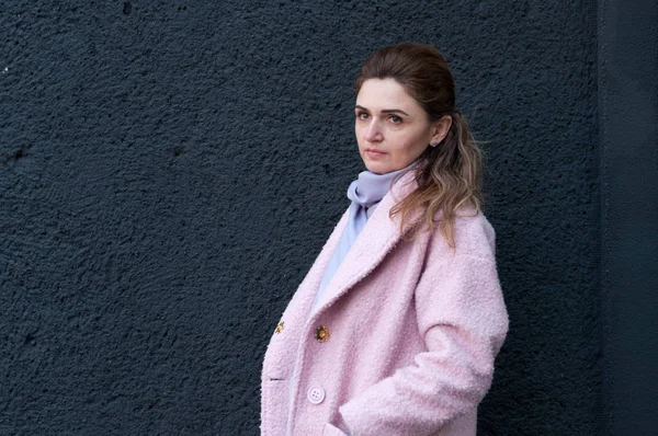 side view of beautiful confident woman in pink coat and scarf looking away and holding hand in pocket on black background with copy space