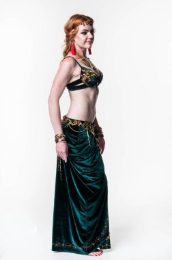 Yeşil kabile dansı kostümlü çekici bir kadın. Üstü ve eteği beyaz arka planda dikiliyor ve başka tarafa bakıyor.