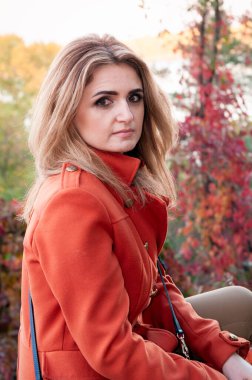 Uzun saçlı, turuncu ceketli, ağaçların önündeki renkli yapraklı kameraya bakan güzel kadının yan görüntüsü.