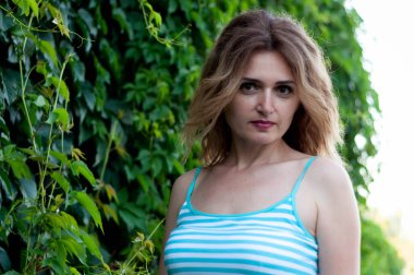 Çekici sarışın kadın, mavi elbiseli, uzun saçlı, yeşil çalıların yanında dikilip kameraya bakıyor.