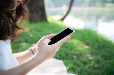 Güzel Asyalı kadınlar ağaçların altında yeşil çimlerin üzerinde oturuyorlar. Akıllı telefon mesajlaşması yapıyorlar.