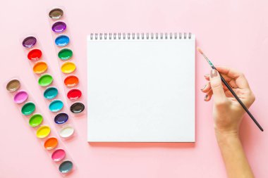 Suluboya paletleri, dizüstü, kadın el tutan boya fırçayaratıcı düz yatıyordu. Pembe pastel arka plan üzerinde Sanatçı işyeri.