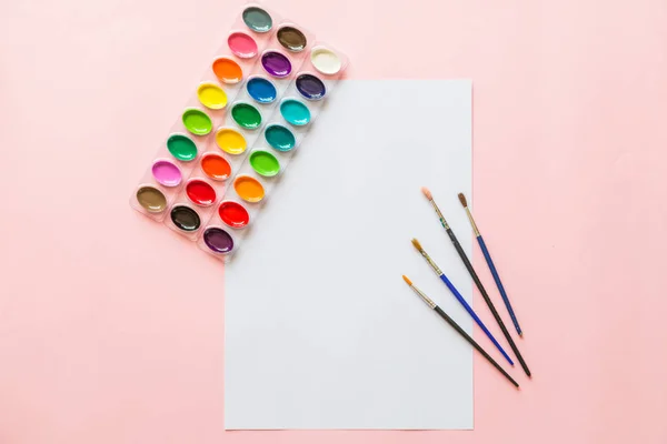 Tvořivé ploché barvy barevných palet, kartáčů na barvy, bílého papíru. Umělecké pracoviště na růžovém pastelu. — Stock fotografie