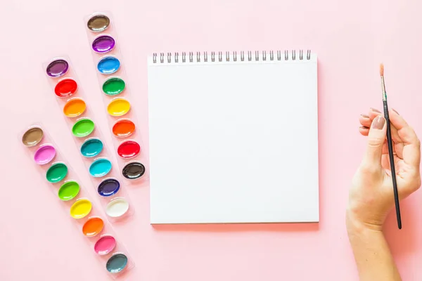 Tvořivé ploché barvy barevných palet, notebooků, ručního držení kartáče. Umělecké pracoviště na růžovém pastelu. — Stock fotografie