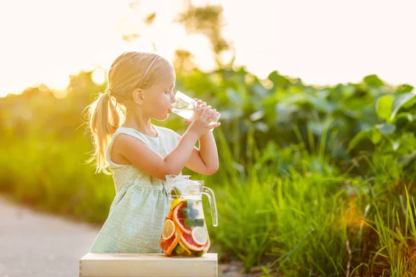 Cute little girl with blonde hair drinking lemonade outdoor. Detox fruit infused flavored water, cocktail in a beverage dispenser with fresh fruits — Stock Photo, Image