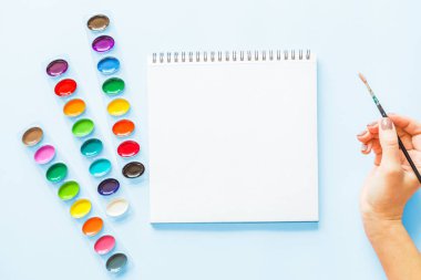 Suluboya paletleri, dizüstü, kadın el tutan boya fırçayaratıcı düz yatıyordu. Mavi pastel arka plan üzerinde sanatçı işyeri.