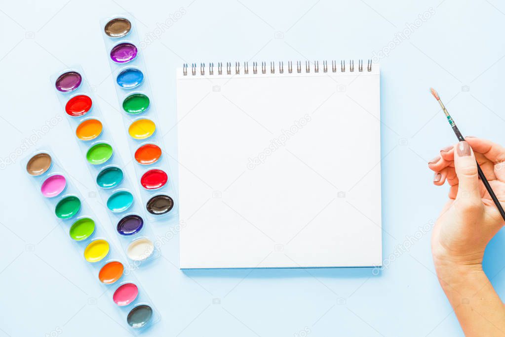 Creative flat lay of watercolor palettes, notebook, female hand holding paint brush. Artist workplace on a blue pastel background.