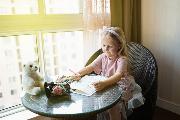 Çocuk kızı annem için kartpostal boyama. Kız masada evde oturan, anne için yanında yatan pembe çiçek. Anneler günü kavramı — Stok fotoğraf