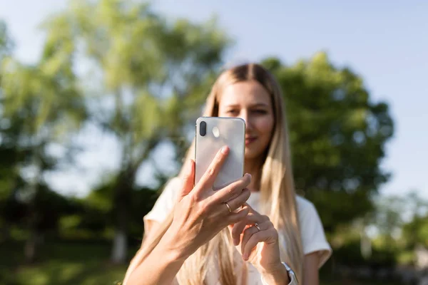 Hermosa mujer joven con cabello rubio utilizando el teléfono móvil al aire libre. Elegante chica haciendo selfie — Foto de Stock