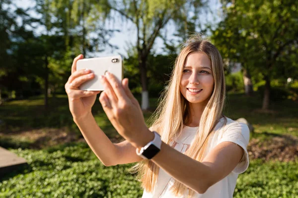 Hermosa mujer joven con cabello rubio utilizando el teléfono móvil al aire libre. Elegante chica haciendo selfie — Foto de Stock